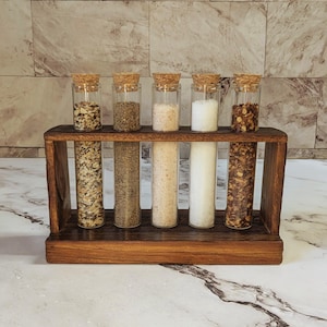 May include: A wooden spice rack with six glass vials, each filled with different spices and topped with a cork. The rack is made of dark wood and sits on a white marble surface. The spices include salt, pepper, and red pepper flakes.