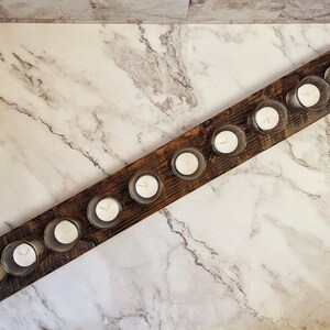 May include: A rustic wooden candle holder with ten clear glass votive holders, each containing a white candle. The candles are arranged in a row on a dark brown, rectangular wooden base. The background is a marble surface.