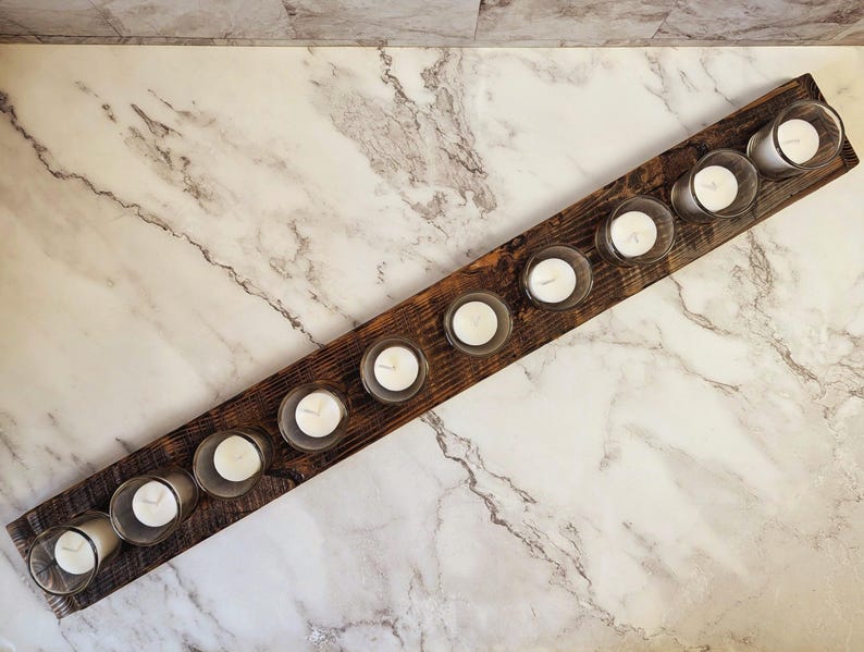May include: A rustic wooden candle holder with ten clear glass votive holders, each containing a white candle. The candles are arranged in a row on a dark brown, rectangular wooden base. The background is a marble surface.