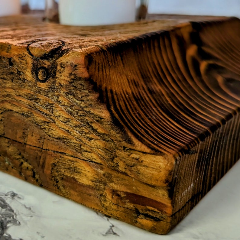 May include: Close-up of a rustic, square wooden block with a warm, amber hue. The wood grain is prominent, featuring dark, contrasting lines and knots. The block appears to be hand-hewn, with a rough-hewn edge. This decorative piece could be used as a candle holder or a small display stand.