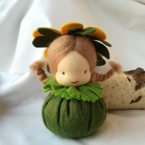 Könnte beinhalten: Eine handgefertigte Filzpuppe mit einem grünen Kleid, einer gelben und grünen Blumenkrone und braunen Haaren. Die Puppe sitzt auf einer weißen Oberfläche.