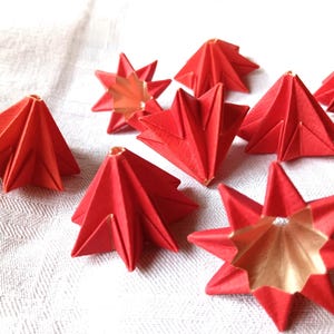 Könnte beinhalten: Eine Sammlung roter Origami-Sterne und sternförmiger Ornamente. Die Papiersterne haben eine goldene Innenseite und sind auf einer weißen, strukturierten Oberfläche angeordnet. Die Origami-Sterne sind eine festliche Dekoration.