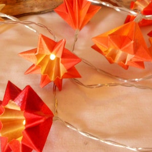 Könnte beinhalten: Eine Lichterkette mit Origami-Papiersternen in Orange- und Rottönen. Jeder Stern beherbergt ein kleines Licht, das ein warmes Leuchten erzeugt. Die Lichter sind auf einem dünnen, silbernen Draht aufgereiht, perfekt für festliche Dekorationen.
