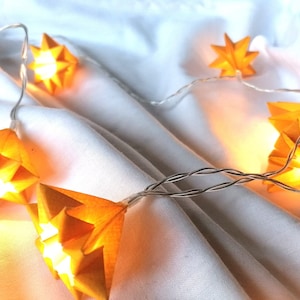 Könnte beinhalten: Eine Lichterkette mit warmgelben sternförmigen Lichtern an einem silbernen Draht, die auf einer weißen Stoffoberfläche liegt. Die Lichter sind eingeschaltet und erzeugen ein sanftes Leuchten. Die Sterne haben ein geometrisches Design.