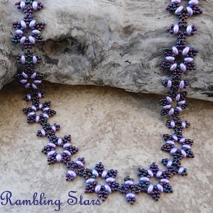 Op de afbeelding: Een kralenketting met een bloemmotief. De ketting heeft lavendelkleurige en iriserende kralen gerangschikt in stervormige patronen. De tekst "Rambling Stars" is zichtbaar linksonder in de afbeelding. De ketting wordt op een neutrale achtergrond gepresenteerd.