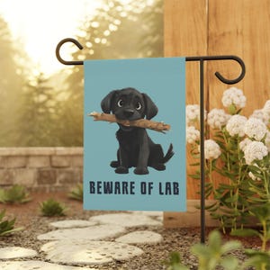 May include: A rectangular garden flag with a light blue background featuring a black Labrador puppy sitting and holding a wooden stick in its mouth. Below the puppy, the text BEWARE OF LAB is printed in dark blue, bold, capital letters.