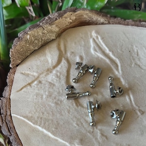 May include: Six small, silver-coloured metal objects with a decorative design. Each object has a cylindrical body, a rounded base, and a small, angled top. The objects are arranged on a light brown, textured surface.