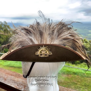 May include: A slouch hat featuring a wide, olive-drab felt brim with a brown leather edge. A gold-toned metal rising sun badge with a crown and text is centred on the front. The hat is adorned with a large plume of long, wispy, brown and black feathers.