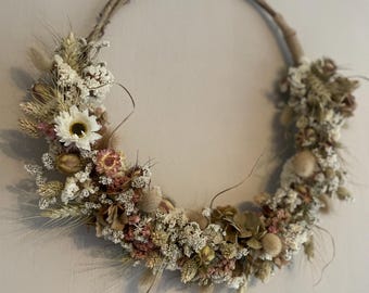 May include: A dried flower wreath with a variety of white, beige, and pink flowers and grasses. The wreath is hanging on a white wall.