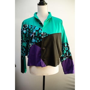 May include: A long-sleeve button-up jacket featuring a color-blocked design in bright teal, solid black, and purple. The garment includes a patterned section with teal and blue geometric shapes on a black background, a collar, and dark buttons.