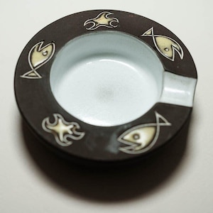 May include: A round, brown ceramic ashtray with a white interior. The rim features stylized fish and wave designs in cream-colored outlines. The ashtray has a built-in rest for cigarettes.