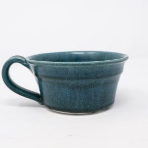 May include: A handmade ceramic mug in a deep teal color. The mug has a rounded body with a handle and a slightly flared rim. The glaze has a speckled appearance, adding texture to the surface. The mug is designed for hot beverages.