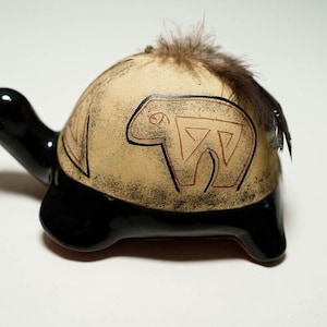 May include: A ceramic turtle figurine with a black shell and a beige top. The top is decorated with a stylized bear design in black and red. The turtle has a small feather on its back.