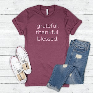 May include: A maroon t-shirt with the words "grateful. thankful. blessed." in white lettering. The shirt is paired with blue jeans and white sneakers with red trim, all arranged on a white wooden surface.
