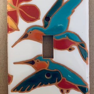 May include: White ceramic light switch cover featuring two teal and orange hummingbirds in flight. The design includes red flowers and is outlined in brown. The switch plate has a single toggle switch opening.