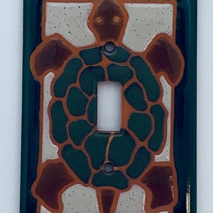 May include: A green ceramic light switch cover with a brown and green turtle design. The turtle has a detailed shell with a white background.