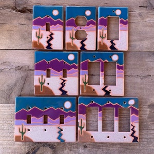 May include: A collection of seven rectangular light switch and outlet covers featuring a desert landscape design. The scene depicts purple mountains, a light purple horizon, a beige desert floor, a green cactus, and a blue sun against a dark blue sky.