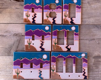May include: A collection of seven rectangular light switch and outlet covers featuring a desert landscape design. The scene depicts purple mountains, a light purple horizon, a beige desert floor, a green cactus, and a blue sun against a dark blue sky.