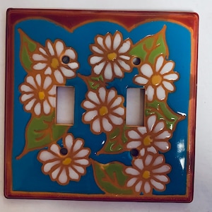 May include: A ceramic light switch cover with a blue background and a white daisy pattern. The cover has a brown border.