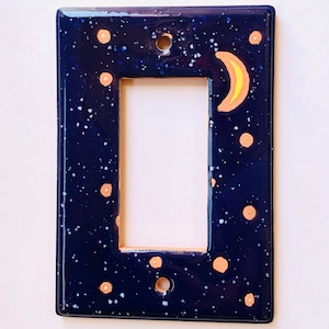 May include: A dark blue ceramic light switch cover with a white and gold speckled pattern. A crescent moon with a yellow interior is painted on the cover.