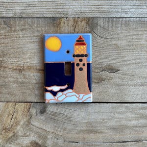 May include: A ceramic light switch cover with a lighthouse, sun, and waves design. The lighthouse is brown with a red roof and black windows. The sun is yellow and the waves are white and blue.