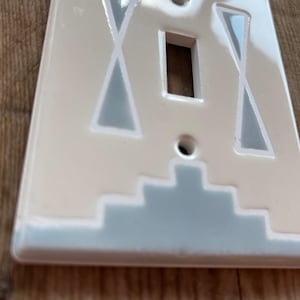 May include: A white ceramic light switch cover with a light blue geometric design. The switch cover has a rectangular shape with a single light switch opening in the center.