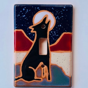 May include: A ceramic light switch cover with a black silhouette of a coyote howling at the moon. The background is a blue night sky with stars and a red and orange mountain range.