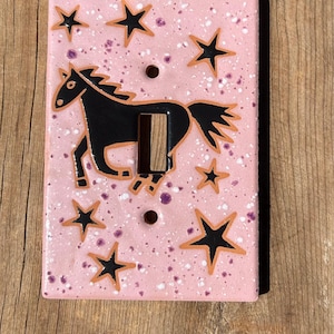 May include: A rectangular light switch plate with a glossy pink finish, featuring a speckled texture with white and purple dots. A black horse silhouette is centered on the plate, surrounded by several black stars with orange outlines.