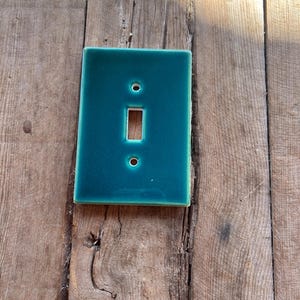 May include: A teal ceramic light switch cover with a glossy finish. The cover has a rectangular shape with a single toggle opening and two screw holes. The switch plate is set against a weathered wooden surface.