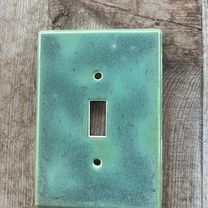 May include: A rectangular light switch cover plate featuring a glossy, mottled sage green glaze with darker green and brown speckling. The plate has a centered rectangular cutout for a toggle switch and two circular screw holes at the top and bottom.