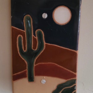May include: A ceramic tile with a desert scene. The tile features a green cactus, a brown and red landscape, and a white moon in a dark blue sky.