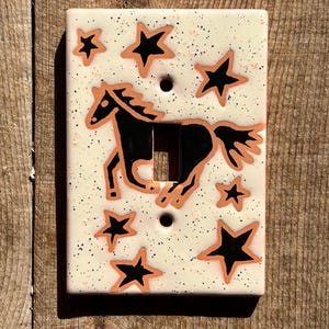 May include: A rectangular ceramic light switch plate with a cream-colored base featuring black speckles. The design includes a black horse silhouette outlined in copper, surrounded by several black stars with copper outlines, and two screw holes.