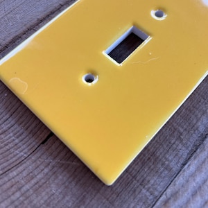 May include: A rectangular light switch wall plate with a glossy, solid yellow finish. The plate features a central rectangular cutout for a toggle switch and two circular screw holes positioned vertically above and below the center opening.
