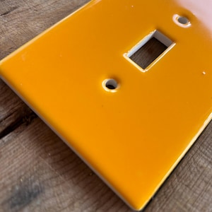 May include: A glossy, bright orange single-gang light switch cover. The cover has a rectangular opening for the switch and two screw holes above and one below. The cover is rectangular with rounded corners.
