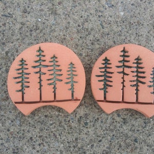 May include: Two terracotta coasters with a black silhouette of a forest of pine trees. The coasters are round with a slight curve on the bottom edge.