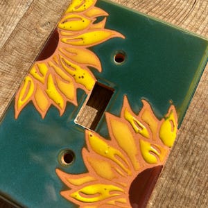 May include: A rectangular ceramic light switch cover plate featuring a glossy dark green background with two hand-painted yellow sunflower designs in opposite corners. The plate includes a central rectangular cutout and two circular screw holes.