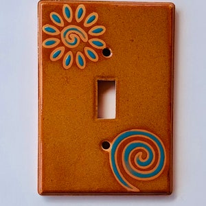 May include: A ceramic light switch cover with a brown background and blue and gold painted designs. The designs include a stylized sun and a spiral.