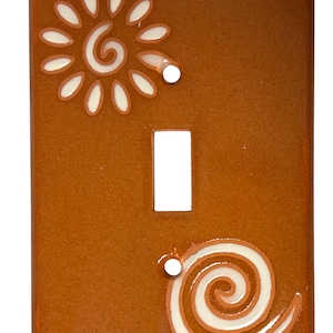 May include: A brown ceramic light switch cover with a white sun and swirl design.