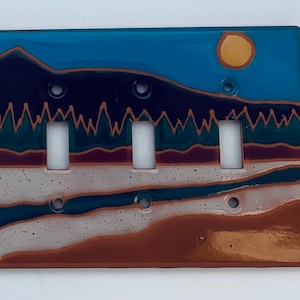 May include: A ceramic light switch cover with a landscape scene. The scene features a blue sky with a yellow sun, a dark blue mountain range, a green forest, and a brown and white riverbed.