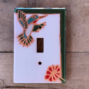 May include: A white ceramic light switch cover with a green border. The cover features a colorful hummingbird and a red flower.
