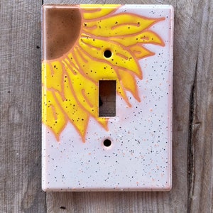 May include: A rectangular light switch cover plate featuring a hand-painted sunflower design. The upper left corner displays a brown center with bright yellow petals extending outward. The background is a speckled off-white with tiny black dots.