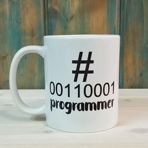 K&ouml;nnte beinhalten: Wei&szlig;er Keramikbecher mit schwarzem Henkel. Der Becher hat ein schwarzes Hashtag-Symbol, Bin&auml;rcode und das Wort "Programmierer" in Schwarz aufgedruckt. Der Hintergrund ist eine Holzoberfl&auml;che.