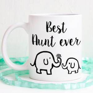 Op de afbeelding: Een witte keramische mok met de zwarte tekst Best Aunt ever in een cursief lettertype. Onder de tekst staat een zwarte lijntekening van een grote olifant die een bloem met zijn slurf vasthoudt, naast een kleine babyolifant.