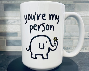 May include: White ceramic mug with the text "you're my person" in black script. A black and white elephant illustration holds a yellow flower. The mug has a large handle.
