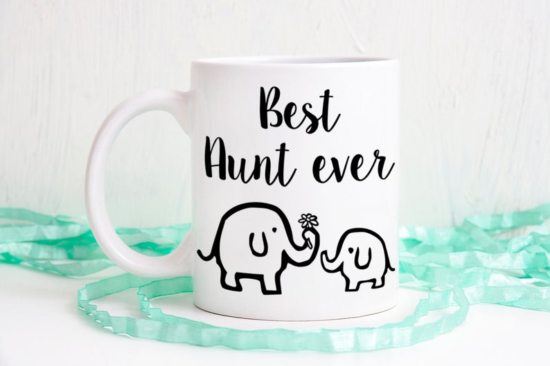 Op de afbeelding: Een witte keramische mok met de zwarte tekst Best Aunt ever in een cursief lettertype. Onder de tekst staat een zwarte lijntekening van een grote olifant die een bloem met zijn slurf vasthoudt, naast een kleine babyolifant.