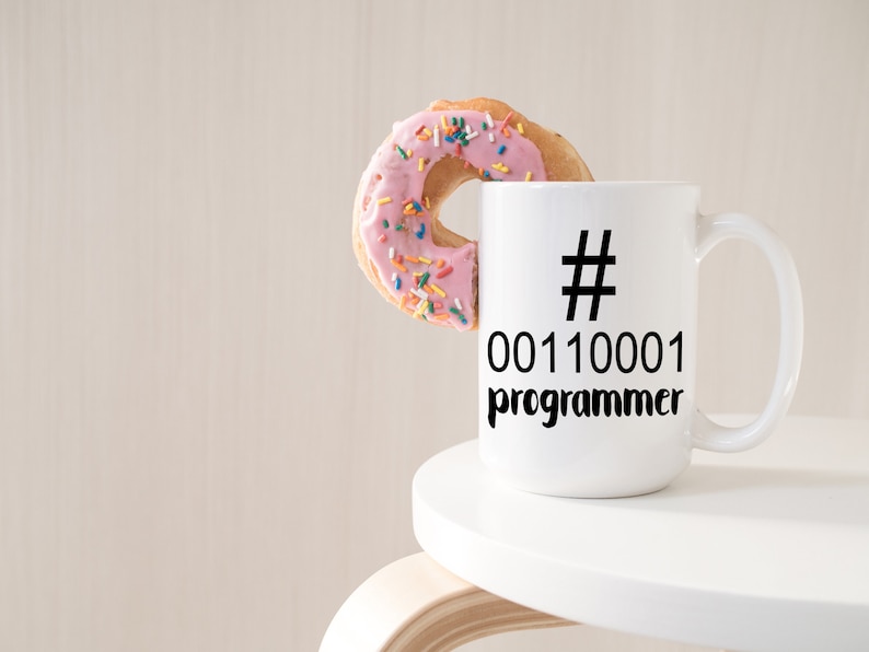 K&ouml;nnte beinhalten: Eine wei&szlig;e Kaffeetasse mit dem Text "# 00110001 programmer" in Schwarz. Ein rosa glasierter Donut mit bunten Streuseln liegt auf dem Tassenrand. Die Tasse steht auf einer wei&szlig;en Oberfl&auml;che.
