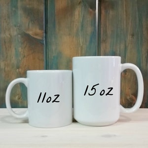May include: Two white ceramic mugs with handles are displayed. The smaller mug is labeled "11oz" and the larger mug is labeled "15oz". The mugs are set against a backdrop of a wooden surface and a blue and brown wooden wall.