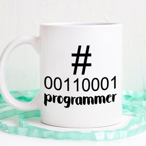 K&ouml;nnte beinhalten: Wei&szlig;er Keramikbecher mit schwarzem Henkel. Der Becher hat ein schwarzes Hashtag-Symbol, Bin&auml;rcode und das Wort "Programmierer" in Schwarz aufgedruckt. Der Becher steht auf einer wei&szlig;en Oberfl&auml;che mit einem hellgr&uuml;nen Band.