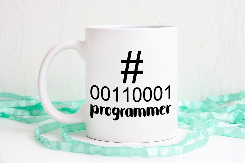 K&ouml;nnte beinhalten: Wei&szlig;er Keramikbecher mit schwarzem Henkel. Der Becher hat ein schwarzes Hashtag-Symbol, Bin&auml;rcode und das Wort "Programmierer" in Schwarz aufgedruckt. Der Becher steht auf einer wei&szlig;en Oberfl&auml;che mit einem hellgr&uuml;nen Band.