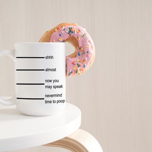 K&ouml;nnte beinhalten: Eine wei&szlig;e Keramiktasse mit schwarzen Linien und dem Text "shhh, fast, jetzt kannst du sprechen, vergiss es Zeit zum Kacken". Ein rosafarbener, glasierter Donut mit bunten Streuseln befindet sich teilweise in der Tasse.